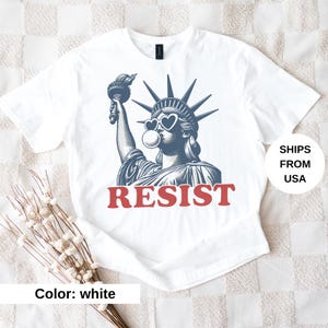 May include: White t-shirt with a graphic of the Statue of Liberty wearing heart-shaped sunglasses and blowing a bubble. The word "RESIST" is printed in red below the image. The shirt is a plain white colour.