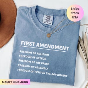 May include: Blue jean colored t-shirt with the text "FIRST AMENDMENT" and a list of freedoms. A gold necklace with a pendant is draped on the shirt. The shirt is labeled "COMFORT COLORS" and the color is "Blue Jean".