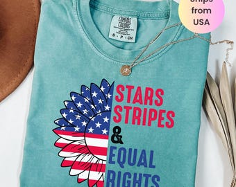 May include: Seafoam green t-shirt with a sunflower design featuring the American flag and the words "STARS, STRIPES & EQUAL RIGHTS." The shirt has a crew neck and short sleeves. A gold necklace with a pendant is on the shirt.