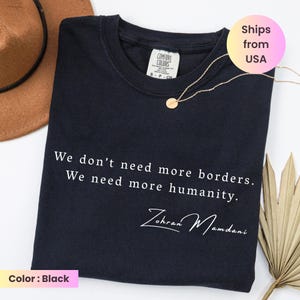 May include: Black Comfort Colors t-shirt with the text "We don't need more borders. We need more humanity." and the signature "Zohran Mamdani" in white. A gold necklace and a brown hat are also visible.