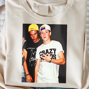 May include: Cream-colored sweatshirt with a printed image of two young men. One man wears a yellow cap, the other a white cap. The man on the right is wearing a white t-shirt with the words "CRAZY KIDS" printed on it.