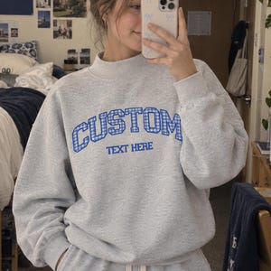 May include: A light grey sweatshirt and matching shorts set. The sweatshirt features a mock neck and the word CUSTOM in blue gingham print, with the text TEXT HERE printed underneath in blue. The shorts have a white drawstring at the waist.