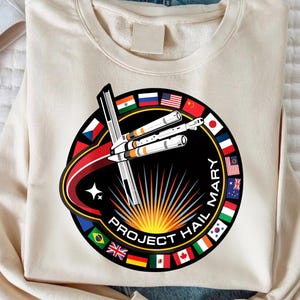 Peut inclure: Sweat-shirt crème avec un motif circulaire représentant une fusée et le texte "PROJECT HAIL MARY". Le motif comprend des drapeaux de différents pays et un soleil stylisé. Le sweat-shirt est fait d'une matière douce.