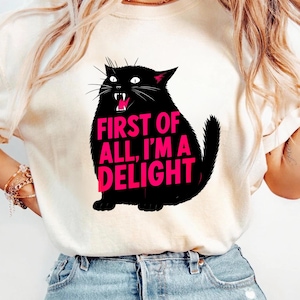 May include: A cream-coloured t-shirt with a black cat graphic and the text "FIRST OF ALL, I'M A DELIGHT" in pink. The cat is shown with an open mouth and sharp teeth, creating a playful and bold design.