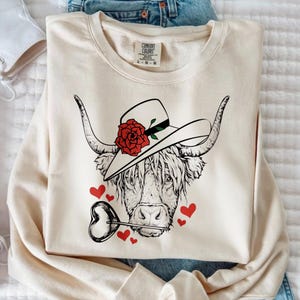 May include: Cream-colored sweatshirt featuring a graphic of a Highland cow wearing a white cowboy hat with a red rose. The cow is holding a heart-shaped lollipop, surrounded by red hearts. The sweatshirt has a Comfort Colors tag.