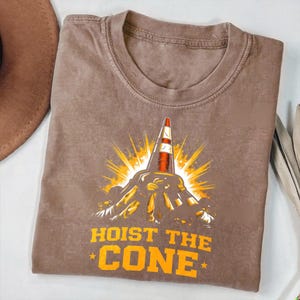 May include: Brown t-shirt with a graphic of a traffic cone, sunburst, and the text "HOIST THE CONE" in orange. The shirt has a crew neck and short sleeves.