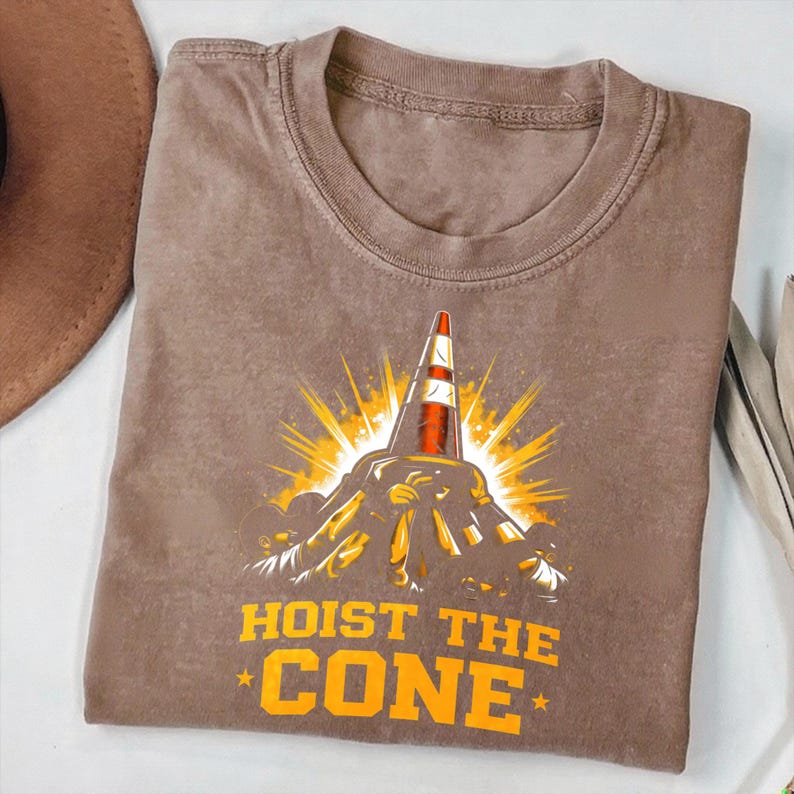 May include: Brown t-shirt with a graphic of a traffic cone, sunburst, and the text "HOIST THE CONE" in orange. The shirt has a crew neck and short sleeves.