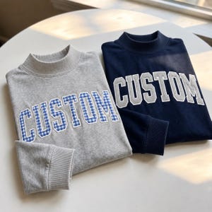 Peut inclure: Deux sweat-shirts à col montant, un gris clair et un bleu marine. Le sweat-shirt gris présente le mot CUSTOM en lettres vichy bleu et blanc. Le sweat-shirt bleu marine présente le mot CUSTOM en lettres bloc gris clair.