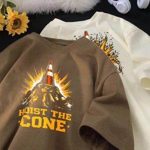 May include: Two short-sleeved t-shirts, one brown and one cream, with a graphic of a traffic cone and the text "HOIST THE CONE" in yellow. The cone is set against a burst of yellow rays.