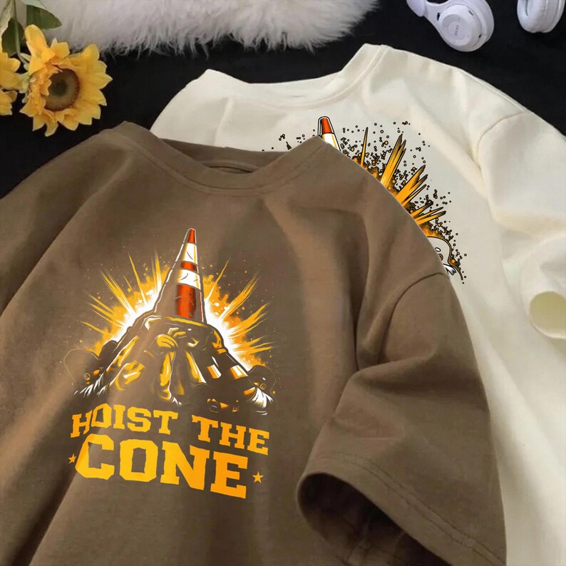 May include: Two short-sleeved t-shirts, one brown and one cream, with a graphic of a traffic cone and the text "HOIST THE CONE" in yellow. The cone is set against a burst of yellow rays.