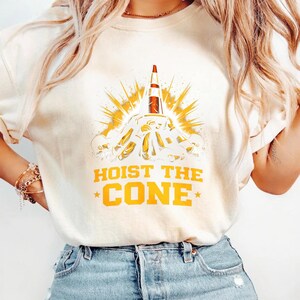 May include: Cream-coloured sweatshirt with a graphic of a traffic cone launching like a rocket, surrounded by yellow and orange rays. The text "HOIST THE CONE" and "CONE" are printed in yellow.