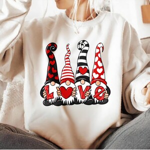 May include: White sweatshirt featuring a graphic of four gnomes holding the letters that spell "LOVE". The gnomes have different patterned hats in red, black, and white, and each gnome holds a letter or a heart. The design is perfect for Valentine's Day.