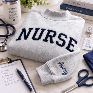 May include: A light grey crewneck sweatshirt featuring the word NURSE in large, dark navy blue block letters across the chest. The left sleeve cuff is embroidered with the name Avery in navy blue script. The fabric has a heathered texture.