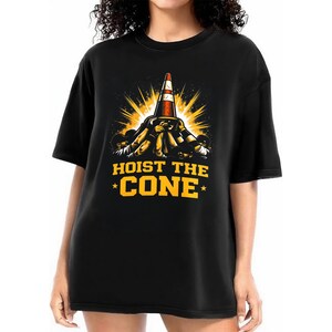 May include: Black t-shirt with a graphic of a traffic cone and the text "HOIST THE CONE" in yellow. The cone is red and white, with a burst of yellow light behind it. The design is centered on the front of the shirt.