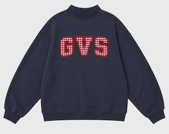 May include: A navy blue long-sleeve sweatshirt featuring a mock neck collar and ribbed cuffs. The chest displays the letters GVS in a red and white gingham pattern with a bold red outline, creating a prominent graphic design on the front.