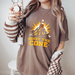 May include: Brown t-shirt featuring a graphic of a traffic cone on a mountain, with yellow rays. The text "HOIST THE CONE" is in yellow, with stars. The shirt is a casual style.