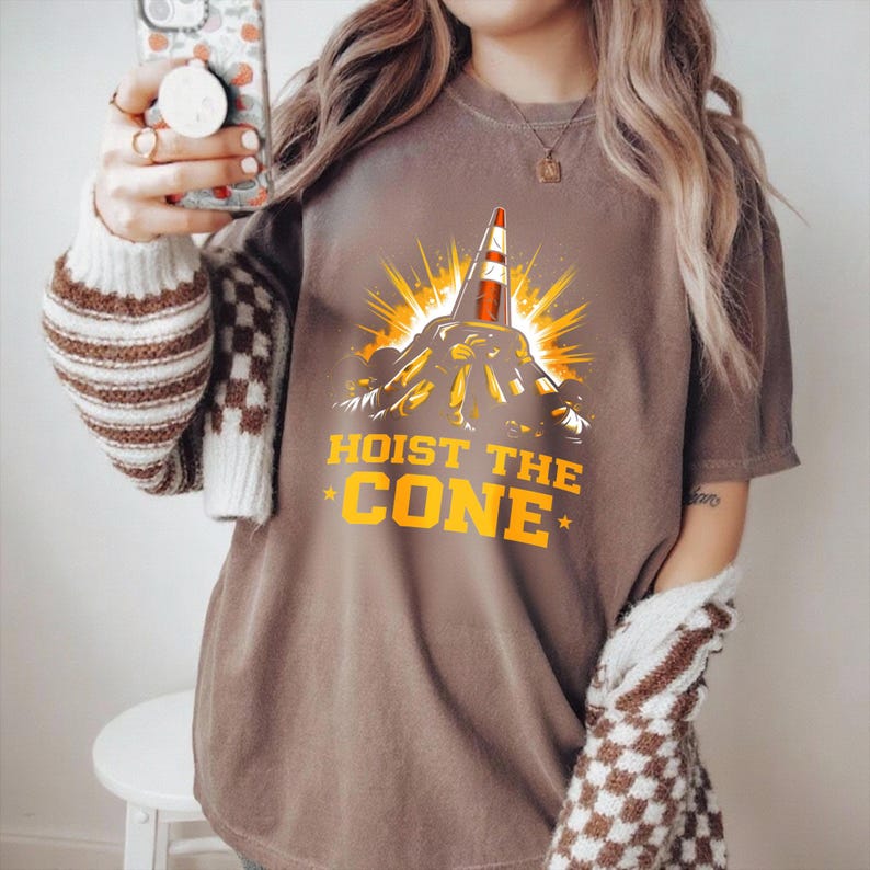 May include: Brown t-shirt featuring a graphic of a traffic cone on a mountain, with yellow rays. The text "HOIST THE CONE" is in yellow, with stars. The shirt is a casual style.