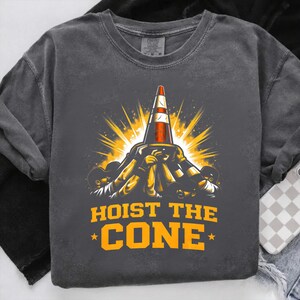 May include: Dark grey crew neck sweatshirt featuring a graphic of a traffic cone being lifted by figures, with a burst of yellow light. The text "HOIST THE CONE" is printed in orange.