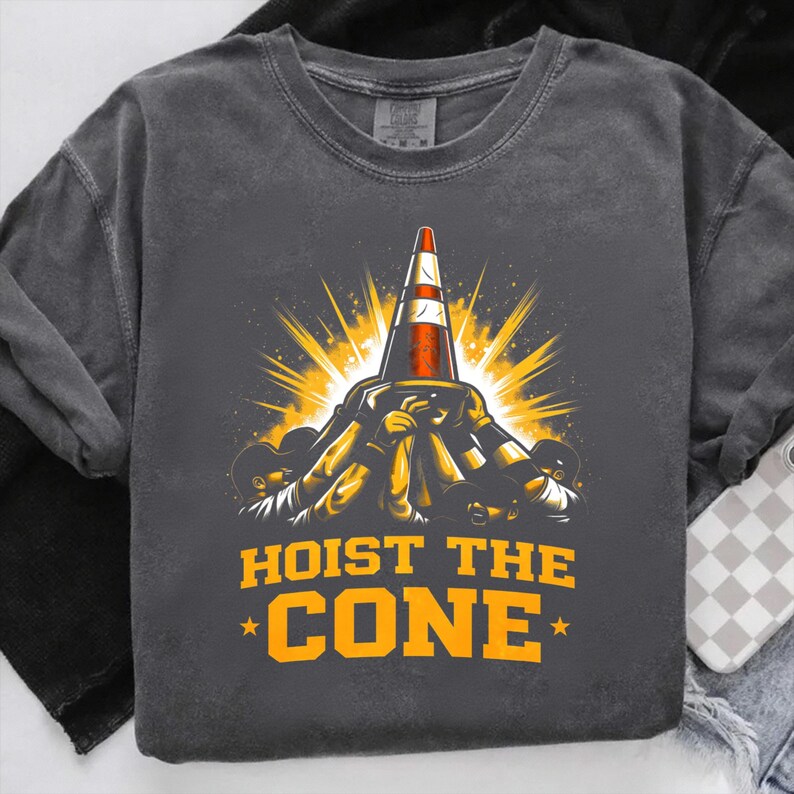 May include: Dark grey crew neck sweatshirt featuring a graphic of a traffic cone being lifted by figures, with a burst of yellow light. The text "HOIST THE CONE" is printed in orange.