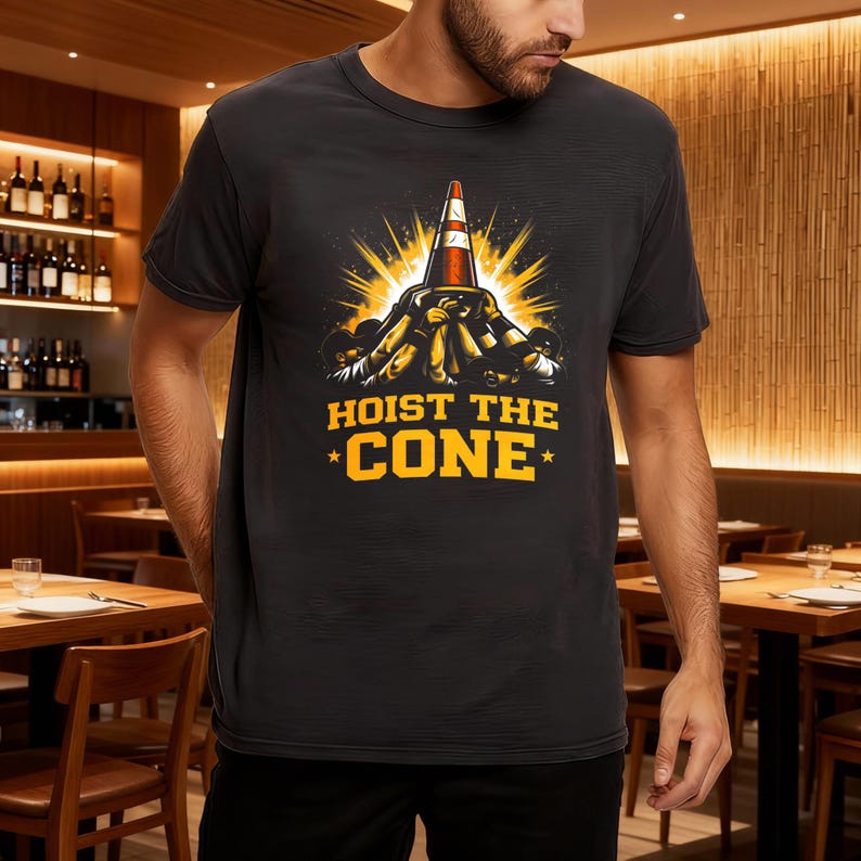 May include: Black t-shirt with a graphic of a traffic cone and the text "HOIST THE CONE" in yellow. The design includes a burst of yellow light behind the cone and figures lifting the cone. The shirt is short-sleeved.