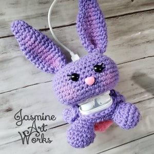 Może przedstawiać: Fioletowy królik szydełkowy z różowymi akcentami trzymający białe etui na bezprzewodowe słuchawki. Królik ma czarne oczy i różowy nos. Etui jest otwarte, a słuchawki są widoczne. Królik siedzi na drewnianej powierzchni.  Jasmine Art Works jest napisane czarnym tekstem w lewym dolnym rogu obrazu.