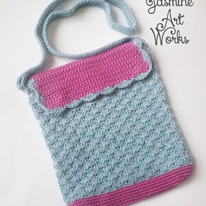 May include: A crocheted shoulder bag with a blue and white patterned body and a pink flap and trim. The bag has a long strap and the text "Jasmine Art Works" is visible in the top right corner of the image.
