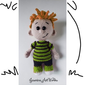Puede incluir: Un niño de juguete de crochet con pelo naranja, una camisa a rayas verdes y negras y pantalones verdes. El juguete está de pie sobre una superficie blanca.