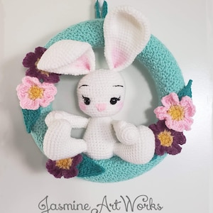 Puede incluir: Una corona de crochet con un conejito blanco sentado en el centro. La corona está decorada con flores rosas, moradas y amarillas.