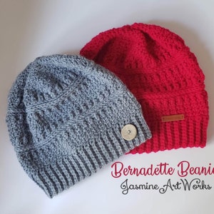May include: Two crocheted beanies, one gray and one red, with a button detail on the gray beanie. The text "Bernadette Beanie Jasmine Art Works" is written below the beanies.