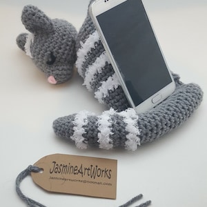 Pode incluir: Suporte de telefone de gato de crochê cinza com listras brancas. O gato está enroscado com o rabo formando um suporte para um smartphone. O telefone não está incluído.