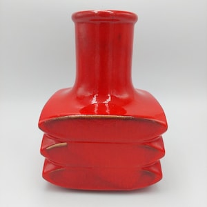 Può includere: Un vaso in ceramica rosso lucido con un design unico e impilato. Il vaso presenta un collo cilindrico e un corpo composto da tre sezioni rettangolari arrotondate impilate. I bordi hanno un sottile bordo marrone. Un pezzo decorativo per la casa.