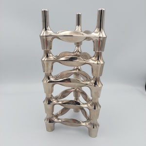 May include: A silver-coloured, geometric wine bottle holder. The holder has a stacked design with open, curved sections and three vertical supports at the top. The base has three feet. The overall design is modern and decorative.