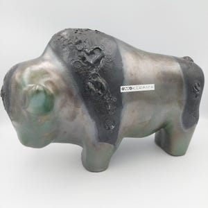 Könnte beinhalten: Eine Keramik-Bisonskulptur in Grau-, Grün- und Schwarztönen. Die Skulptur hat eine strukturierte Oberfläche mit einem dunklen Streifen auf dem Rücken. Der Text "OTTO KERAMIK" befindet sich auf einem weißen Etikett an der Seite. Der Bison steht.