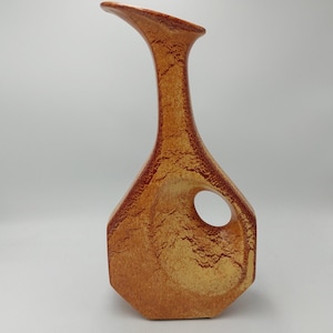 Könnte beinhalten: Eine hohe, strukturierte Keramikvase in Braun- und Beigetönen. Die Vase hat eine einzigartige, asymmetrische Form mit einem gebogenen Ausguss und einer kreisförmigen Öffnung an der Seite. Die Oberfläche hat ein gesprenkeltes Aussehen.