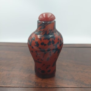 Op de afbeelding: Kleine, decoratieve fles met een rood-zwart gespikkeld patroon. De fles heeft een afgeronde vorm met een bijpassende rode stopper. De fles staat op een houten oppervlak.
