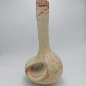 Puede incluir: Un jarrón de cerámica alto, color crema, con una base bulbosa y un cuello delgado. El jarrón presenta un agujero único a través de la base y un patrón texturizado y moteado en tonos marrones y rojos. El borde superior es de un marrón rojizo más oscuro.