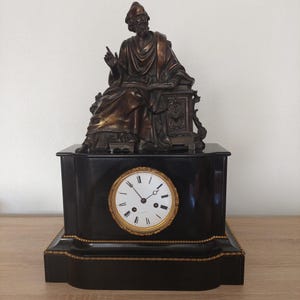 May include: Antique black mantel clock with a bronze statue of a seated figure. The clock has a white clock face with black Roman numerals and gold trim. The statue is detailed with draped clothing and a book.