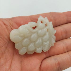 May include: A carved off-white jade pendant shaped like a cluster of grapes with detailed leaves and vines. The stone has a smooth, polished surface with translucent qualities and intricate relief work depicting individual fruit shapes and foliage.