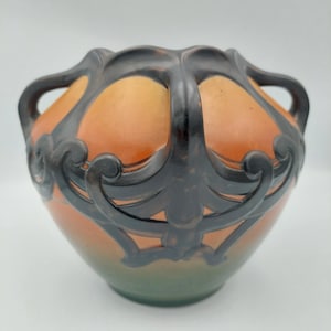 May include: A rounded ceramic vase featuring a gradient color scheme that transitions from a deep green base to a warm orange upper section. The vessel is adorned with a dark, raised, scroll-like overlay that forms integrated handles.