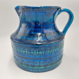 May include: A blue ceramic jug with a handle and spout. The base features a geometric pattern in teal and blue. The jug is glazed, giving it a glossy finish. The overall design is vintage.