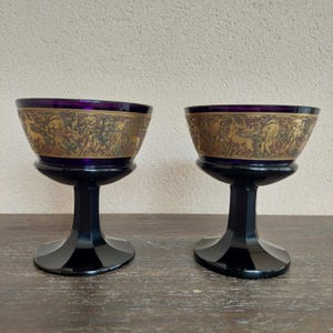 Może przedstawiać: Dwa antyczne kielichy z głęboką fioletową czarką i złotym pasem z bogatym wzorem reliefowym. Kielichy mają czarną nóżkę i podstawę i są prezentowane na drewnianej powierzchni.