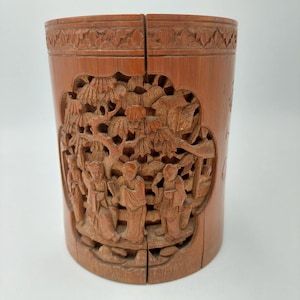 Peut inclure: Un pot à pinceaux cylindrique en bambou brun clair avec des sculptures en relief complexes. Les sculptures représentent des personnages dans un paysage, entourés d'arbres et d'éléments architecturaux. Les bords supérieur et inférieur présentent des bordures décoratives sculptées.