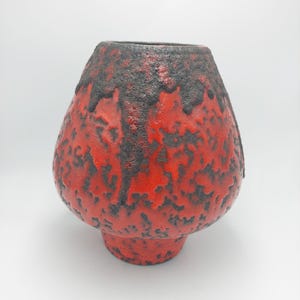 Può includere: Un vaso in ceramica rosso e nero con una superficie strutturata, simile a quella vulcanica. Il vaso ha un corpo bulboso e una base corta e rotonda. La parte superiore del vaso è di colore nero più scuro.
