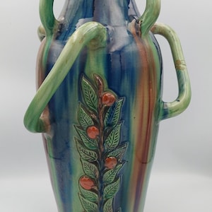 Könnte beinhalten: Eine hohe Keramikvase mit farbenfrohem, glasiertem Finish. Die Vase hat ein Design aus grünen, blauen und braunen Farbtönen mit einem Relief eines belaubten Zweigs mit roten Beeren. Sie hat zwei große, gebogene Griffe und einen kleineren Griff auf jeder Seite.