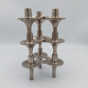 May include: A silver-toned metal candle holder with a unique, interconnected design. It features multiple candle holders at varying heights, with a three-legged base. The overall aesthetic is vintage and decorative.