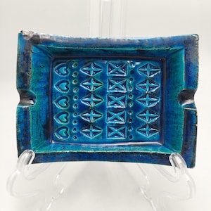 May include: Rectangular ceramic ashtray in shades of blue and turquoise. The ashtray features a raised geometric pattern of crosses, squares, and hearts. It has two built-in rests for cigarettes. The ashtray is approximately 18 cm long and 13 cm wide.