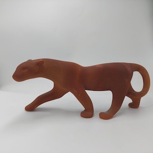 Könnte beinhalten: Eine terrakottafarbene Pantherfigur. Die Skulptur zeigt die Großkatze in einer Laufpose, mit eingerolltem Schwanz. Die Figur besteht aus einem festen Material und hat eine glatte, matte Oberfläche. Die Form des Panthers ist stromlinienförmig und stilisiert.