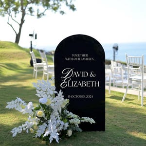 Op de afbeelding: Een zwart trouwbord met een gewelfde bovenkant, met de namen "David & Elizabeth" en de datum "15 oktober 2024" in witte letters. Het bord is versierd met witte bloemen en groen, geplaatst op een grasveld met witte stoelen op de achtergrond.