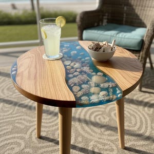 May include: A round side table with a distinctive design. The table has a river of blue resin with embedded seashells. It has a light wood finish and three wooden legs. A glass of lemonade sits on the table. The table is approximately 60 cm in diameter.