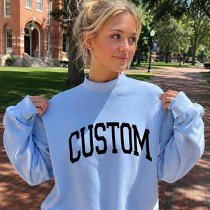 May include: A light blue crewneck sweatshirt featuring a mock neck collar and long sleeves. The word CUSTOM is printed across the chest in a bold, black, arched serif font. The garment is constructed with ribbed cuffs and a ribbed hemline.